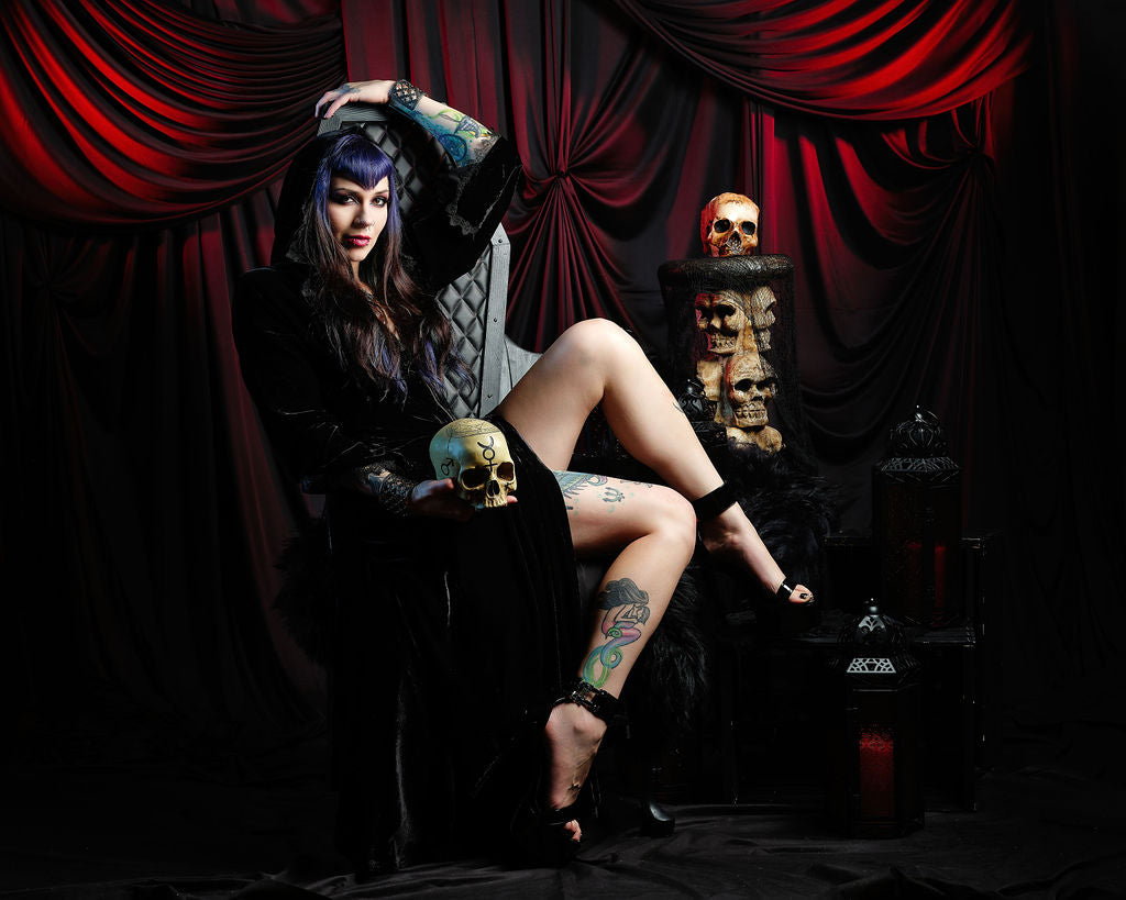 A gothic-style fashion portrait of a woman with purple hair and tattoos, wearing a black dress and sitting on a black throne chair surrounded by skulls and red draped curtains.